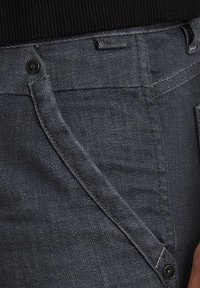 Graue Denim-Jeans mit einer Nahaufnahme der Tasche, sichtbaren Nähten und einem schwarzen Knopfdetail am Bund.