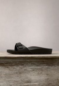 Sandale noire à enfiler avec une bride ajustable à boucle unique et une semelle texturée posée sur une surface en bois.