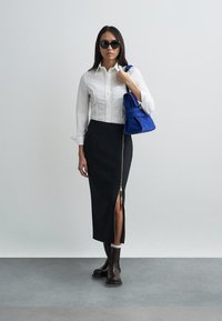 Λευκό πουκάμισο με κουμπιά, μαύρη midi φούστα με φερμουάρ στο πλάι και σκούρα καφέ μποτάκια αστραγάλου. Τσάντα από μπλε σουέτ προσθέτει χρωματική αντίθεση.