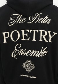 Melnas kapuces džemperis ar lieliem baltajiem uzrakstiem: "The Delta," "POETRY," un "Ensemble," ar dekoratīvu ziedu grafiku un "DON'T WASTE CULTURE" apakšā.
