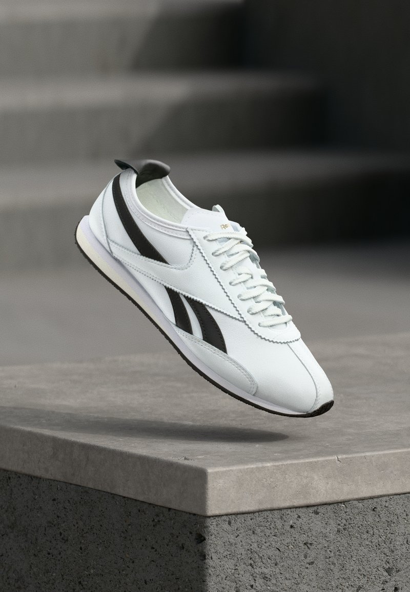 Adidași albi cu dungi negre flotând deasupra unei margini de beton, pe un fundal estompat de o scară gri.