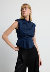 Camiseta sin mangas azul marino con una silueta estructurada, cierre de botones en la parte delantera y cuello puntiagudo. Tejido suave y brillante con detalles fruncidos en el busto.