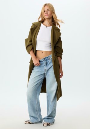 Un trench en vert olive sur un haut blanc court, associé à un jean large bleu clair et des baskets à motifs. Textures douces et coupe décontractée.