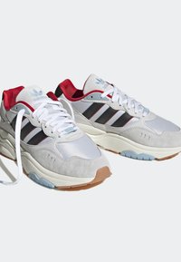 Ett par vita och grå Adidas-sneakers med svarta ränder, röd innersula, vita skosnören och beige sulor på en vit bakgrund.