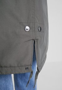 Szara parka z zapięciami na srebrne guziki, rozciętym dołem i regulowanymi sznurkami na dole, ukazująca gładką fakturę tkaniny.