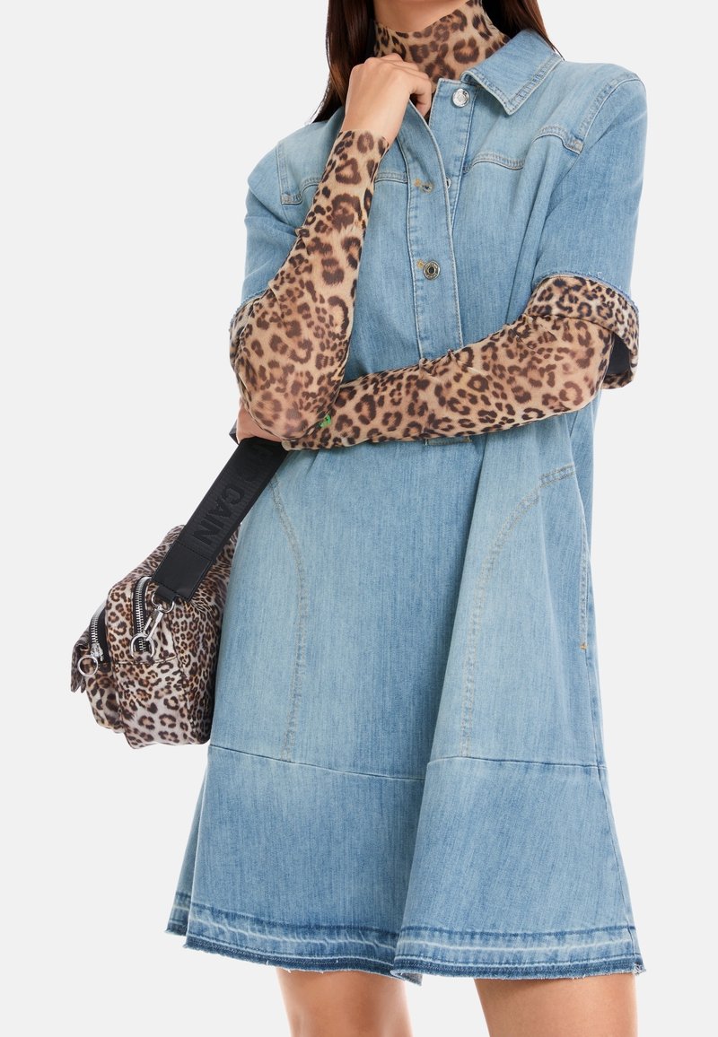 Femme portant une robe en denim bleu clair par-dessus un top à manches longues imprimé léopard, portant un sac imprimé léopard avec une sangle noire sur fond blanc.