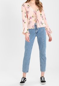 Blusa rosa chiaro con stampa floreale, scollo a V e dettaglio con fiocco frontale. Presenta maniche a campana ed è abbinata a jeans blu chiaro e sneakers nere.