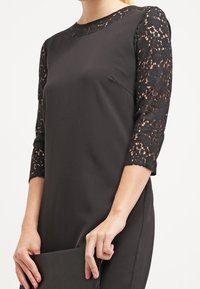 Vestido negro con mangas de encaje tres cuartos, escote redondo y tejido suave. La modelo sostiene un bolso negro de mano, resaltando la simplicidad del vestido.