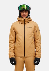 Giacca da sci color camoscio con tasca con zip e cappuccio regolabile, realizzata in materiale impermeabile. Abbinata a guanti neri e casco da sci.