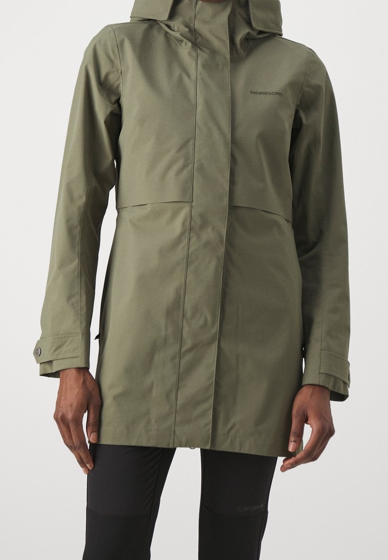 Personne portant une veste imperméable vert olive avec une capuche et des leggings noirs, debout devant un fond clair uni.