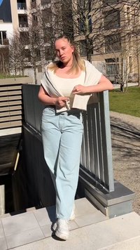 Cardigã cropped bege claro sobre uma blusa ajustada branca, combinado com jeans soltos azuis claros e sapatilhas brancas. Cenário ao ar livre com vegetação.