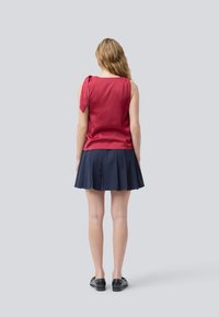 Femme aux longs cheveux ondulés portant un haut sans manches rouge avec un lien à l'épaule, une jupe plissée bleu marine et des chaussures noires à enfiler, se tenant de dos.