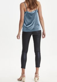 Kvinna klädd i en blå sammetscamisole, svarta åtsittande läderbyxor och beige högklackade sandaler, stående med ryggen vänd framåt.