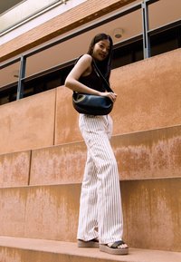 Schwarze Handtasche, ärmelloses Oberteil, weiße gestreifte weite Hose und schwarze Plateausandalen, auf Treppen mit einem strukturierten orangefarbenen Wandhintergrund.