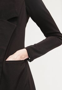 Vêtement noir à manches longues présentant un design à panneaux structurés et une poche latérale, fabriqué à partir d'un tissu lisse et léger.