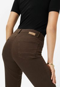 Jean en denim marron avec une coupe ajustée, des poches arrière et une étiquette beige sur la ceinture. Porté avec une chemise noire.