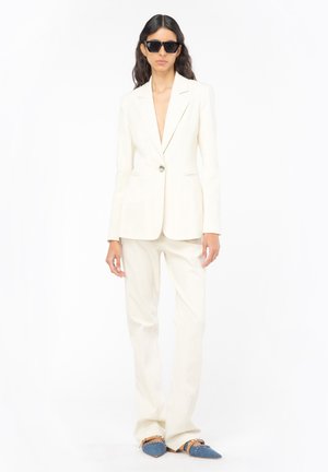 Blazer crema con un bottone singolo e revers a punta, abbinato a pantaloni coordinati. Accentato con scarpe flat blu a punta con decorazioni.