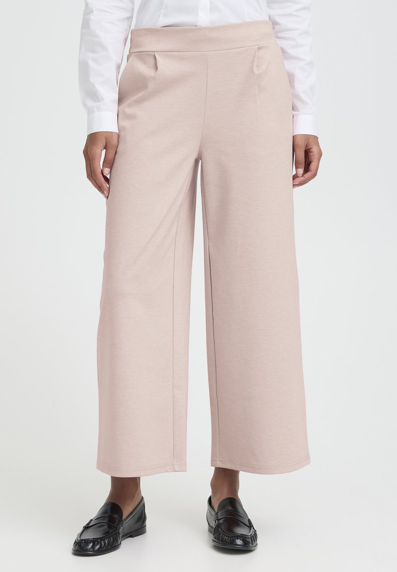 Mujer con pantalones beige de pierna ancha, camisa blanca de manga larga y mocasines negros sobre un fondo liso.
