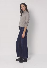 Donna in piedi di profilo che indossa un dolcevita grigio chiaro, jeans larghi a gamba larga di colore scuro e stivaletti neri, su uno sfondo bianco neutro.