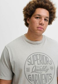 Hellgraues T-Shirt mit rundem Grafiklogo in Dunkelgrau, das "Superior Quality Garments" liest. Kurze Ärmel, aus weichem Baumwollmaterial, entspannte Passform.