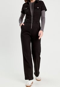 Černý džínový overal s krátkými rukávy, náprsními kapsami a předním zipem. Nošený přes dlouhý šedý top, doplněný bílými teniskami.
