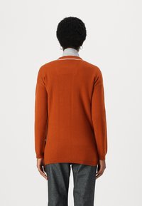 Orange Strickpullover mit langen Ärmeln, Rundhalsausschnitt und geripptem Saum. Kombiniert mit grauen, strukturierten Hosen, von hinten betrachtet.