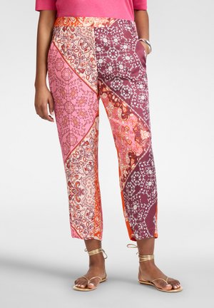 Vrouw die kleurrijke, gebloemde cropped broek draagt met roze, oranje en paarse paisley patronen, gecombineerd met gouden sandalen met enkelriem.