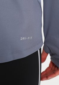 Ljusgrå prestandatopp med meshtextur, med "DRI-FIT"-logotyp; matchad med svarta byxor som har vita sidostrimmor.