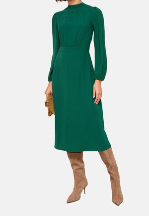 Femme portant une robe midi verte à manches longues et col haut, tenant une pochette marron, associée à des bottes à talons en daim beige montant jusqu'aux genoux.