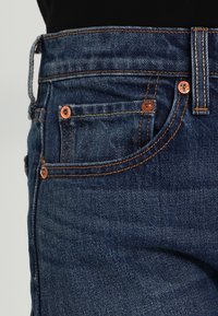 Primer plano de un bolsillo de jeans de mezclilla azul con remaches de cobre y costuras naranjas sobre un fondo negro.