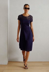Marinedress med blonde korte ermer, kneelengde og glatt tekstur. Kombinert med svarte stroppesandaler, på et lyst tregulv.
