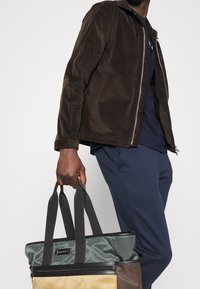 Brown corduroy jacket with front zipper, blue shirt underneath, and gray-brown tote bag with black straps and accent hardware.