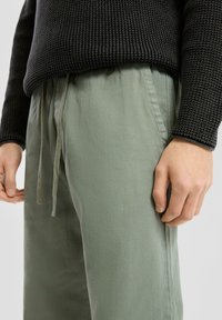 Pantalons vert clair, coupe décontractée avec une texture. Dotés d'une taille à cordon de serrage et de poches latérales. Associés à un pull en tricot noir.