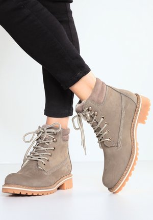 Personne portant des bottines taupe à lacets avec col rembourré et semelles en caoutchouc naturel, associées à un pantalon noir ajusté.