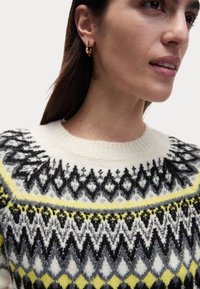 Krämfärgad tröja med svarta, grå och gula geometriska mönster; ribbad hals. Gyllene hoopsmycke synligt i modellens högra öra.