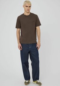 T-shirt en coton marron à manches courtes, associé à un jean en denim bleu avec des coutures blanches. Les chaussures comprennent des baskets vertes et jaunes.