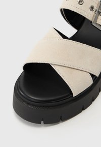 Beige suede sandals featuring crisscross straps, adjustable buckle, and a thick black platform sole with a textured surface.