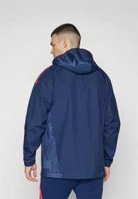 Chaqueta impermeable de color marino con capucha, puños elásticos y acentos rojos en los hombros; cuenta con un acabado suave y mate, y un fit relajado.