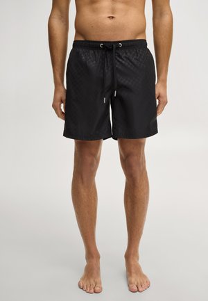 Bloot bovenlichaam persoon die zwarte badshorts met patroon en trekkoord draagt, blootsvoets staand op een neutrale achtergrond.