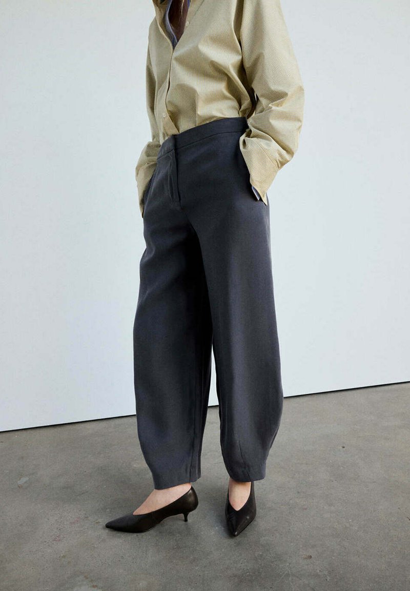 Personne portant une chemise beige avec les mains dans les poches, un pantalon ample foncé et des chaussures noires à talons bas, debout sur un sol en béton.