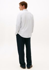 Camicia bianca a maniche lunghe con un motivo testurizzato, abbinata a pantaloni blu navy, vista dal retro, evidenziando l'orlo curvo della camicia.