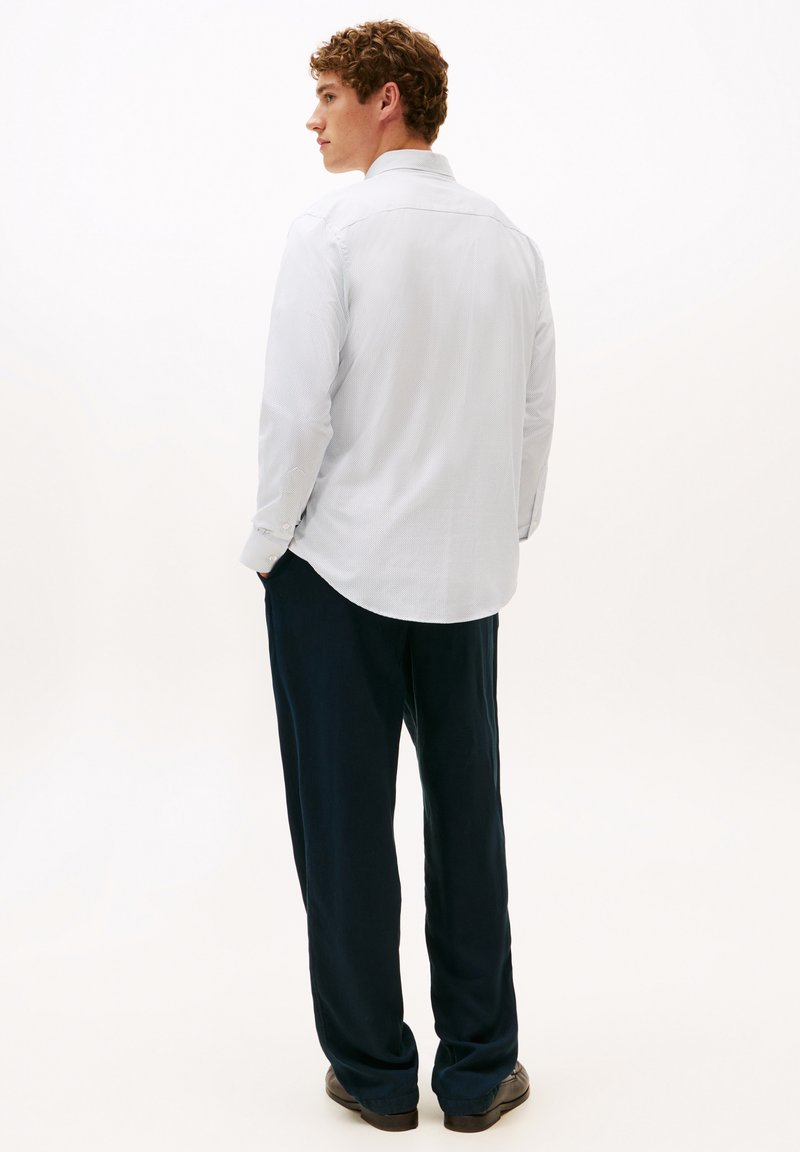 Witte lange mouwen blouse met een gestructureerd patroon, gecombineerd met marineblauwe broek, gezien van achteren, waarbij de afgeronde zoom van de blouse wordt benadrukt.