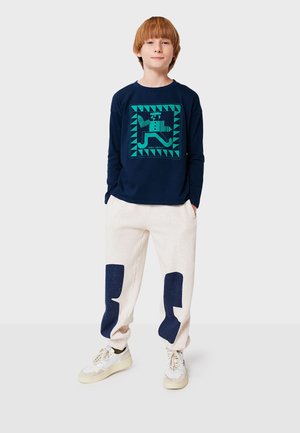 Chemise à manches longues marine avec un design géométrique vert, assortie à un pantalon de jogging beige clair avec des patchs bleu foncé. Portée avec des baskets claires.