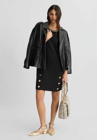 Abito nero con dettagli di bottoni dorati, indossato sotto una giacca di pelle nera. Accompagnato da una borsa in pelle di serpente e scarpe flat dorate.