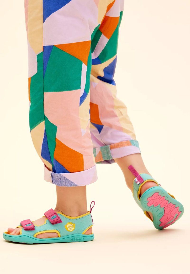 Sandalias ajustables de color azul claro con acentos en rosa y amarillo. Cuentan con una suela acolchada y un diseño de punta abierta. Pantalones con un patrón geométrico colorido.