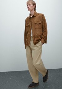 Brown suede shirt jacket with two chest pockets, paired with beige high-waisted pants and dark shoes on a textured gray floor.
