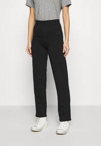 Schwarze, hochgeschnittene Hose mit geradem Bein, aus weichem Material, kombiniert mit weißen Sneakers und einem grauen T-Shirt.
