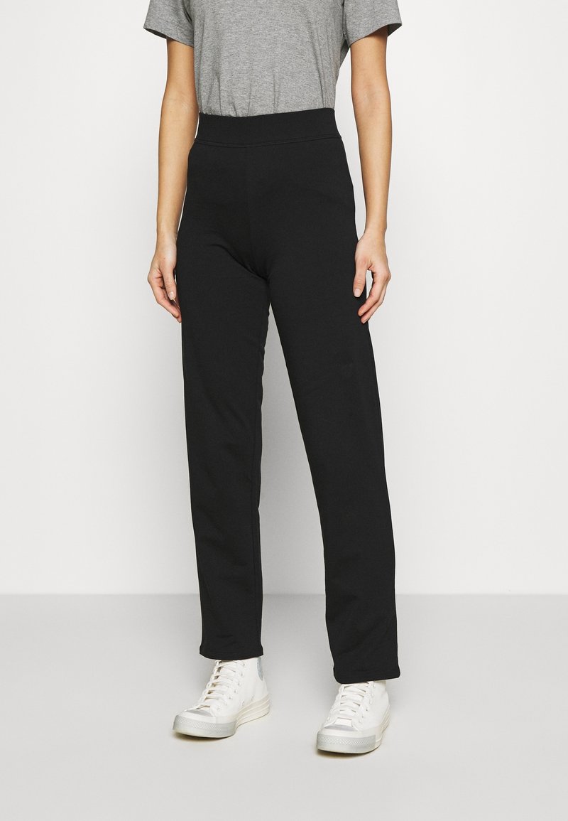 Schwarze, hochgeschnittene Hose mit geradem Bein, aus weichem Material, kombiniert mit weißen Sneakers und einem grauen T-Shirt.
