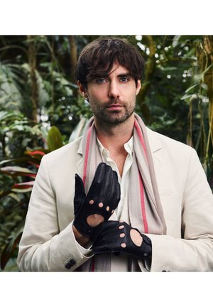 Man in beige colbert en sjaal past zwarte leren handschoenen aan, staande voor een achtergrond van groene tropische bladeren.