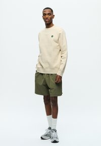 Beige sweatshirt with small green logo, paired with loose olive green shorts, white crew socks, and grey athletic shoes on a plain background.